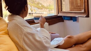 A person in a white bathrobe sitting by a window with a view of a mountainous landscape. - Complejo Turístico Rural La Garganta (Álora)