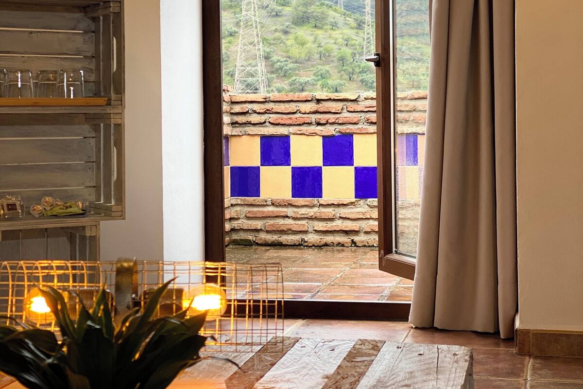 A room with a view of a mountainous landscape through a glass door, a potted plant, a wooden floor, and a shelf with glassware.