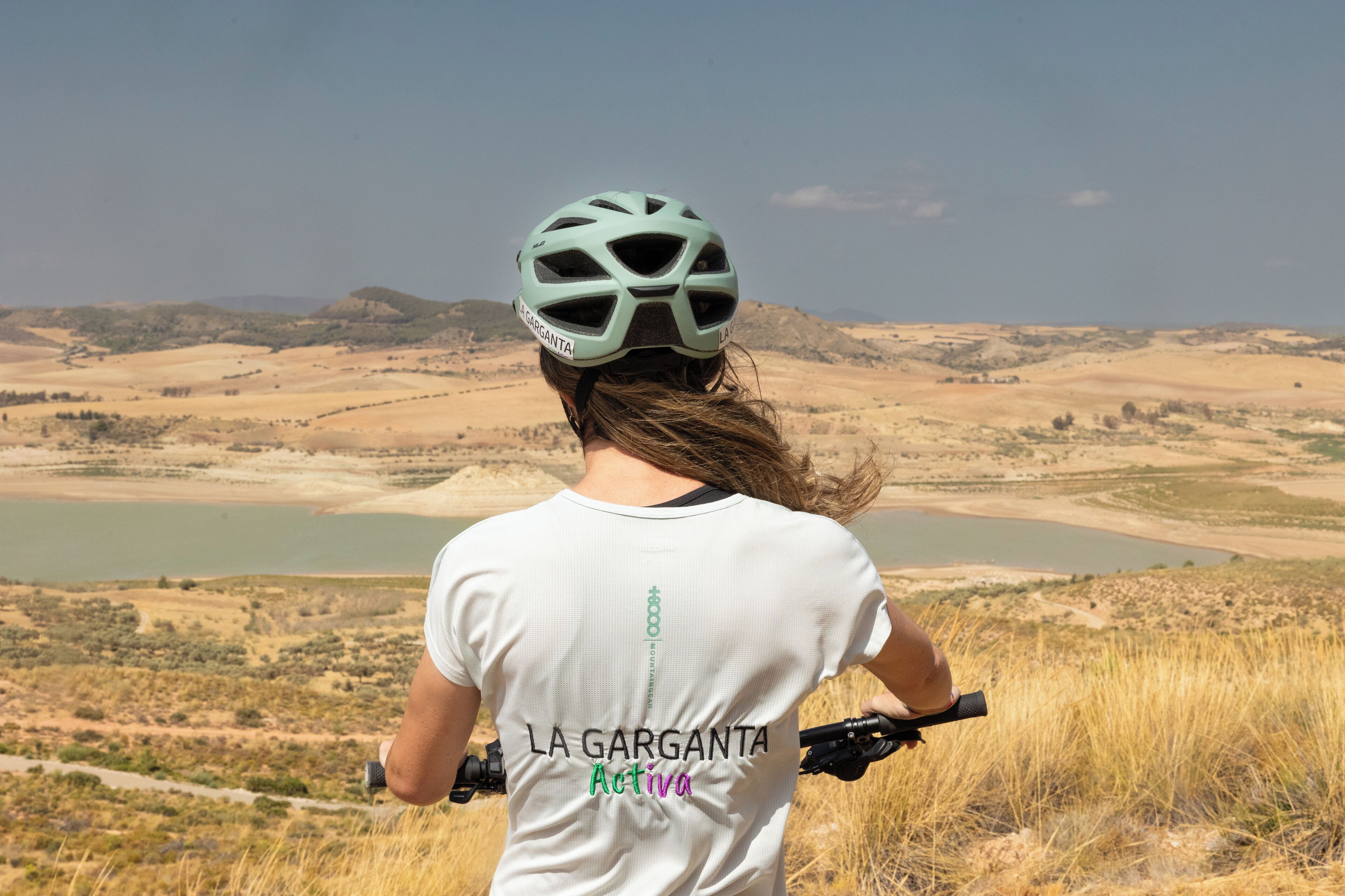 A person wearing a helmet and a white t-shirt with "LA GARGANTA Activa" printed on the back, standing in a natural landscape with hills and a body of water in the background.