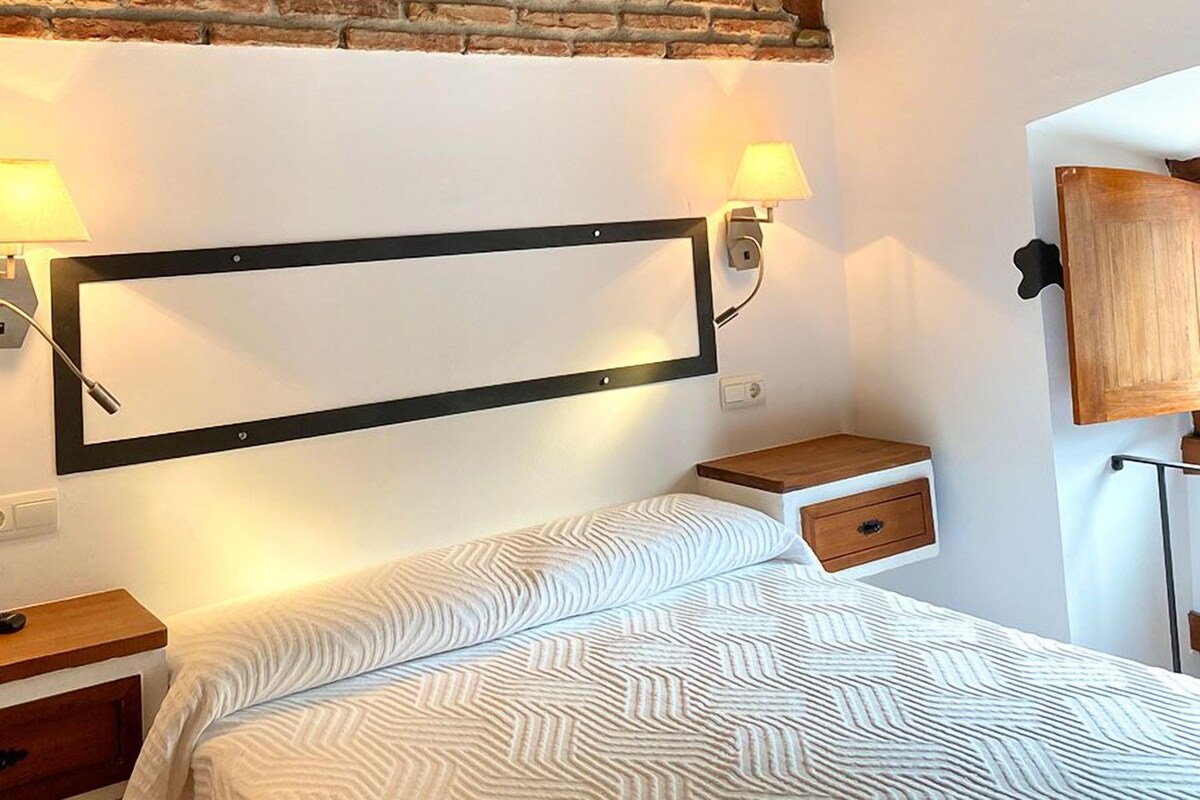 A bedroom with a bed, bedside tables, a wall-mounted light, and a mirror.