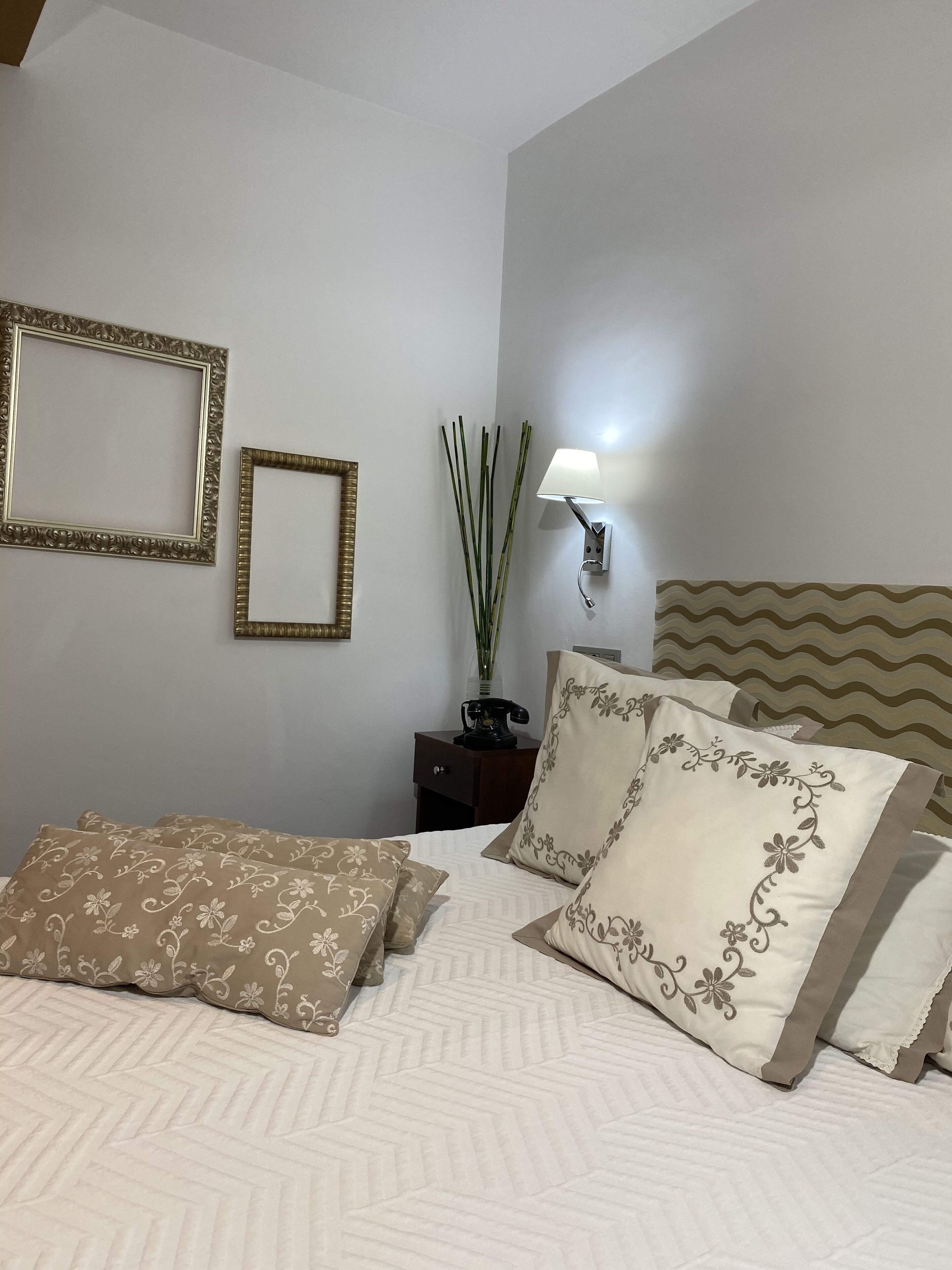 Un letto rifatto con cuscini decorati, una lampada a parete e due quadri appesi.