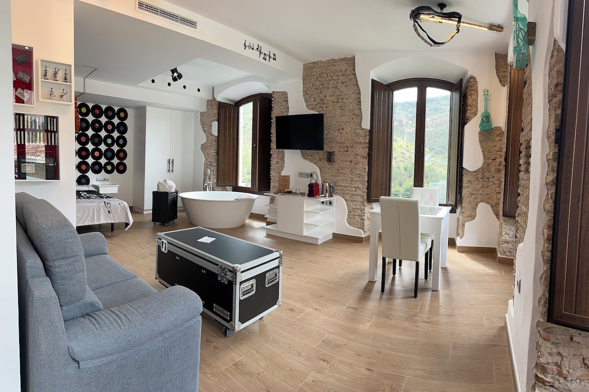 A modern living room with a grey sofa, a black portable storage unit, a white bathtub, and a dining area with white chairs.