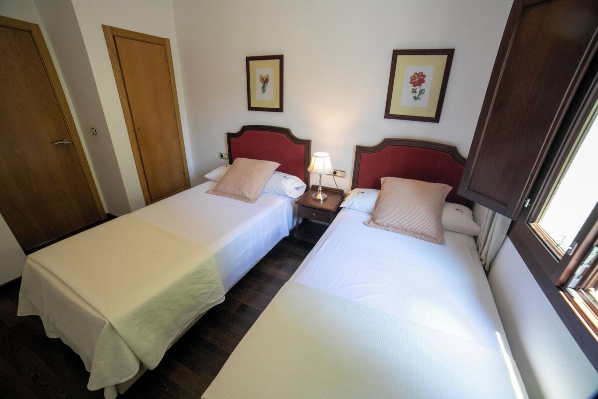 A hotel room with two single beds, wooden doors, framed floral artwork, a bedside table with a lamp, and a window.