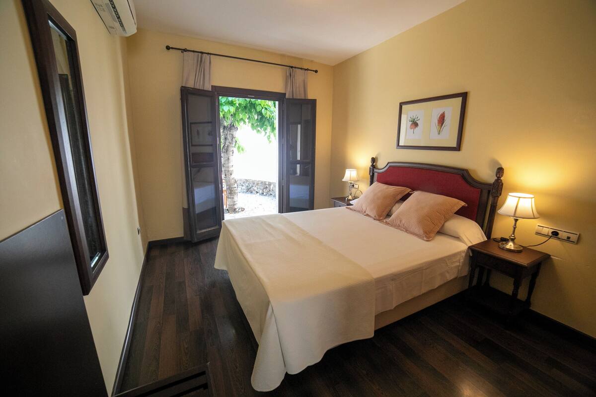 A bedroom with a large bed, a nightstand with a lamp, a window with curtains, and a door leading to an outdoor area.