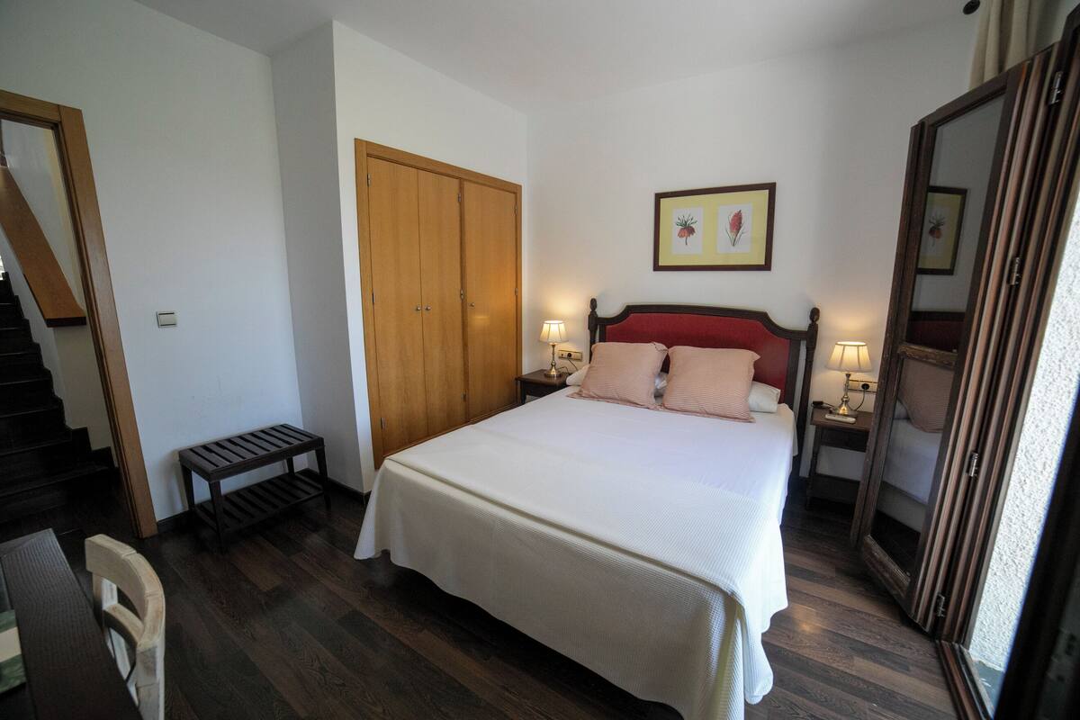 A bedroom with a bed, wooden wardrobe, bedside tables with lamps, a bench, and a mirror.