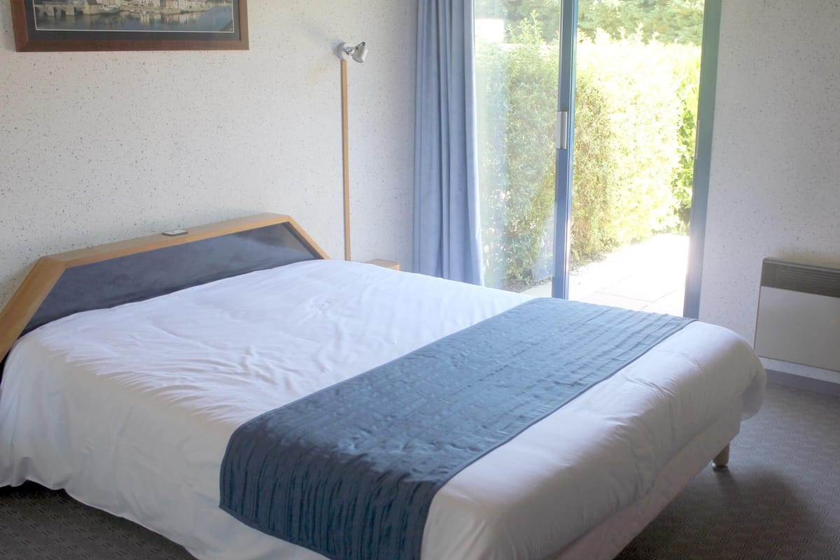A neatly made bed with a headboard, a bedside table, and a window with curtains.