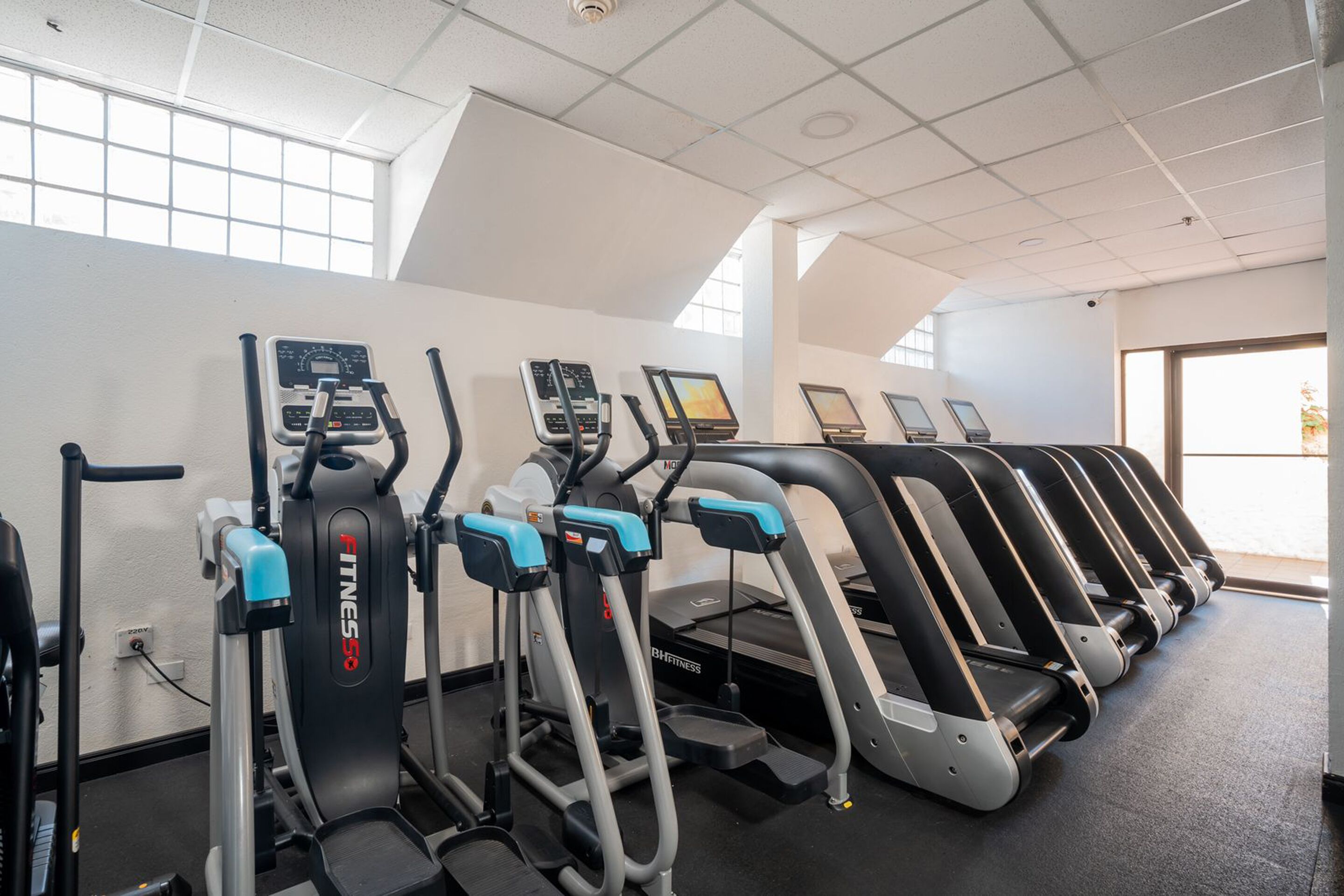 Una hilera de cintas de correr y máquinas elípticas en un gimnasio con ventanas amplias y techo alto.