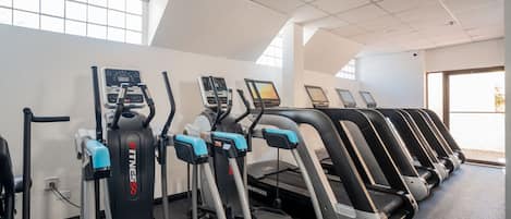 Una hilera de cintas de correr y máquinas elípticas en un gimnasio con ventanas amplias y techo alto.