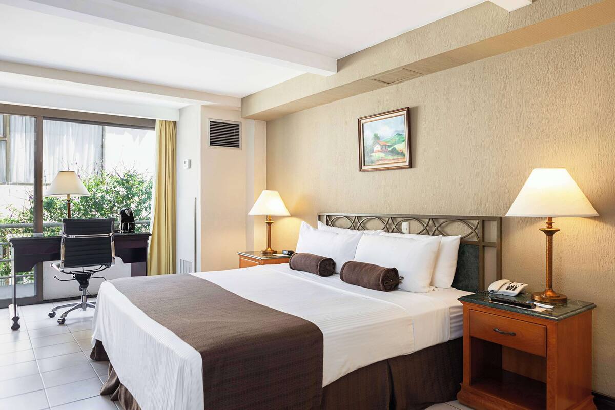 A hotel room with a large bed, two bedside tables with lamps, a desk with a chair, and a window with curtains.