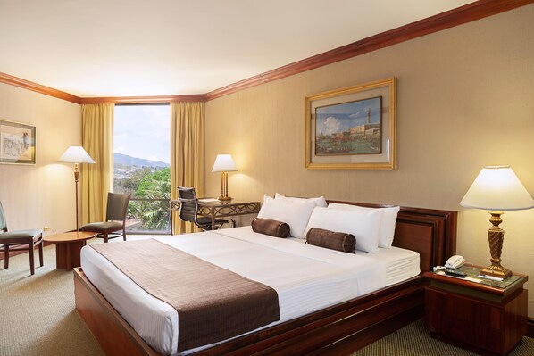 A hotel room with a large bed, two bedside lamps, a desk with a chair, and a painting on the wall.