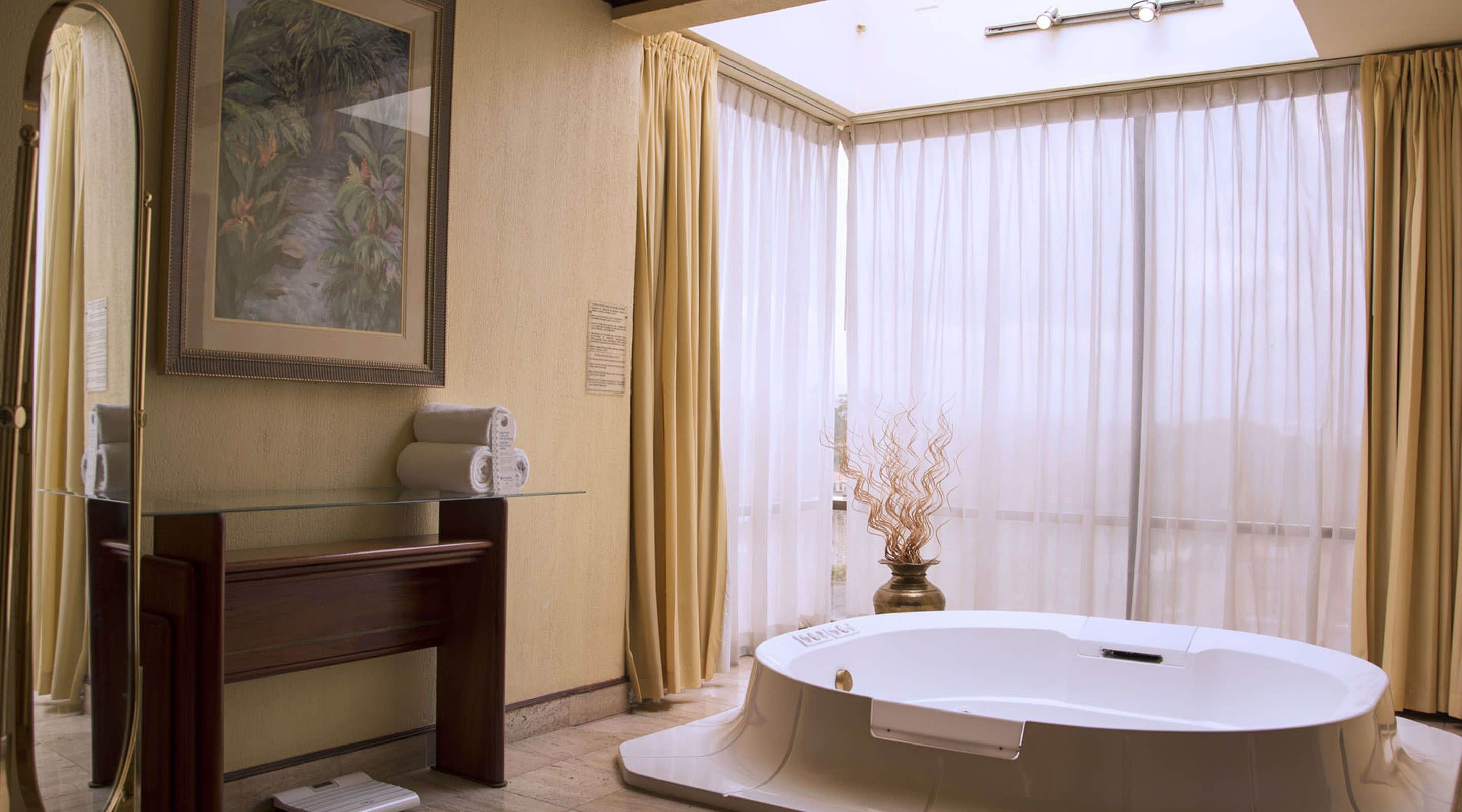 A bathroom with a glass vanity, a large mirror, a framed painting, a bathtub, and sheer curtains.
