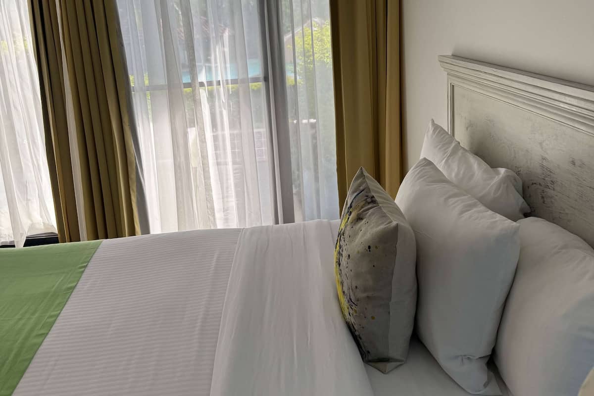 A neatly made bed with white linens and a green bedspread, a wooden headboard, and sheer curtains allowing natural light.