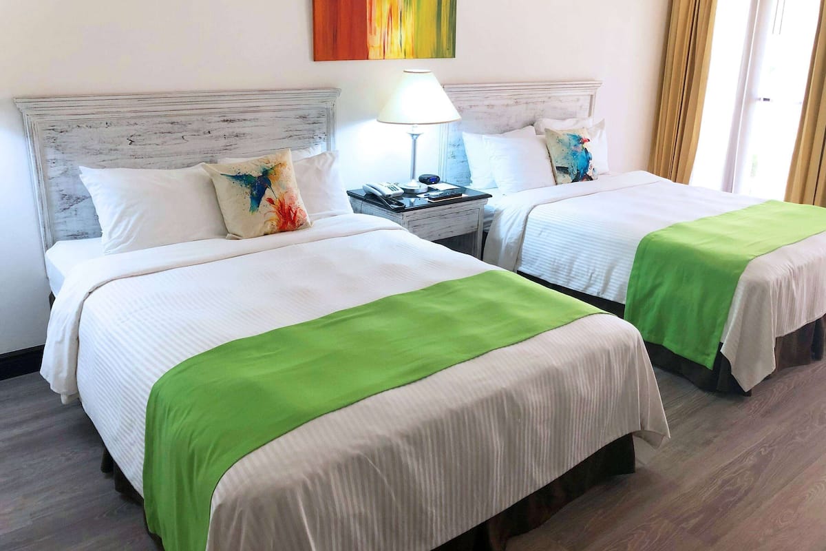 A hotel room with two beds, each with a green and white bedspread, a white headboard, and a bedside table with a lamp.