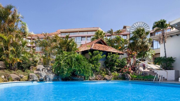 Una piscina con una pequeña cascada, rodeada de plantas tropicales y palmeras, con un edificio con techo de tejas rojas al fondo.