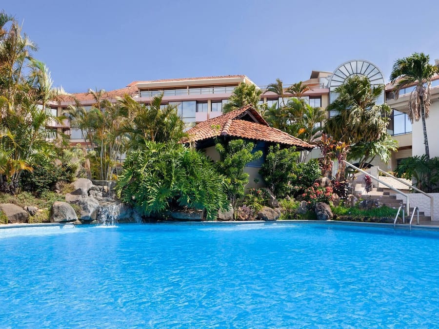 Piscina com uma pequena cascata, rodeada por plantas tropicais e palmeiras, com um edifício com telhado de terracota ao fundo.