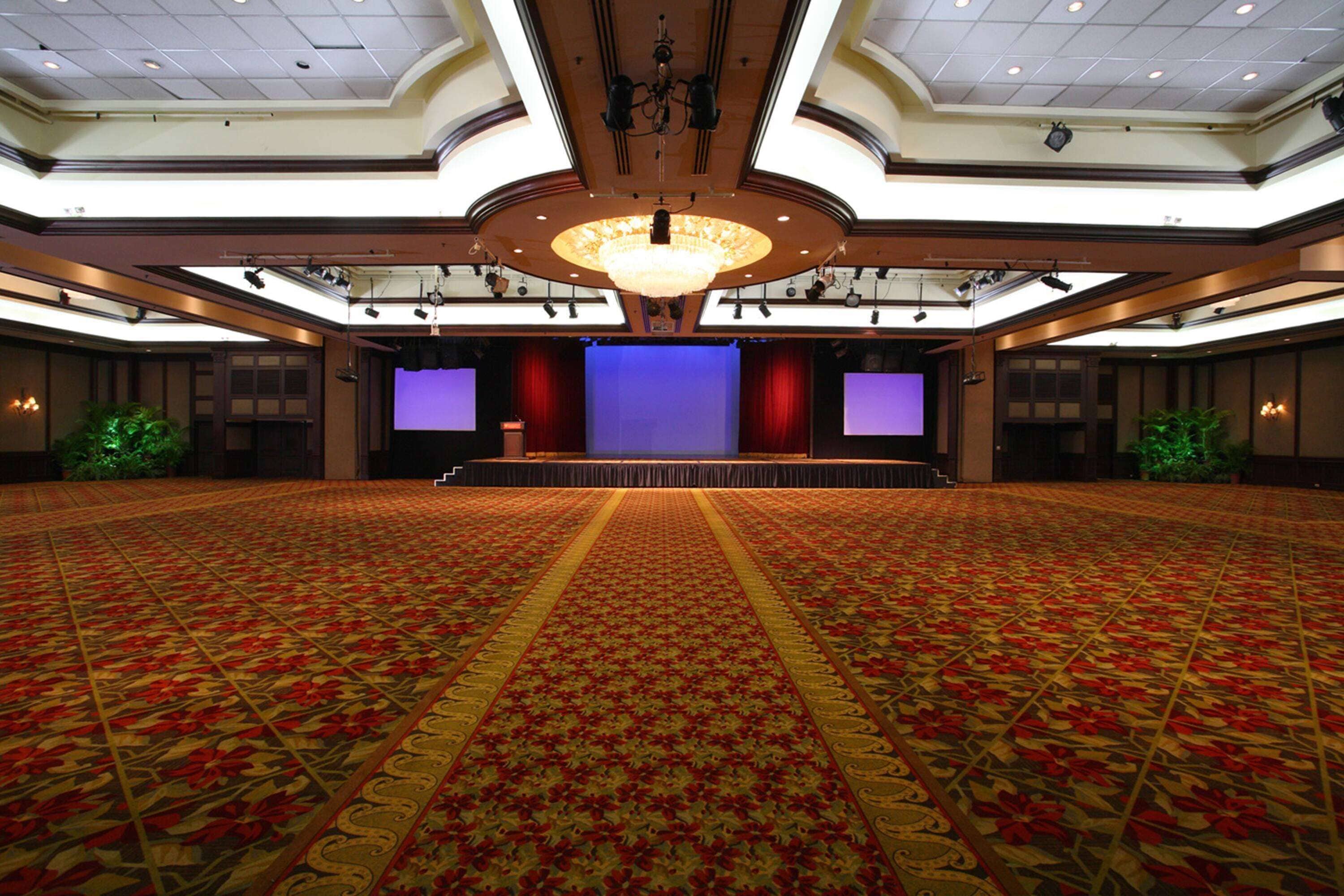 Une grande salle de bal dotée d’un tapis à motifs, d’une scène centrale avec des rideaux et d’un lustre.