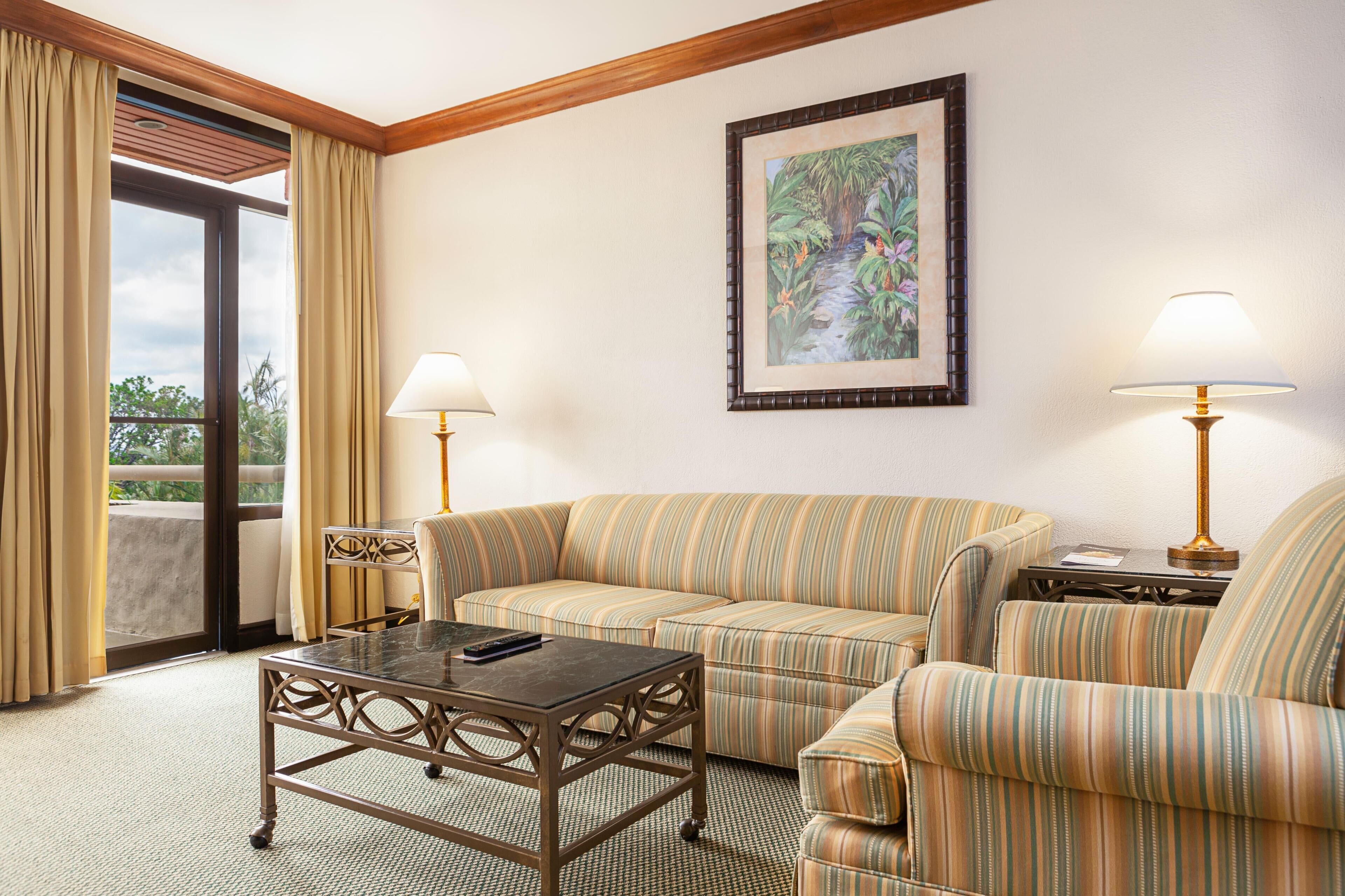 A room with a sofa, armchair, table, lamps, and a framed picture on the wall.