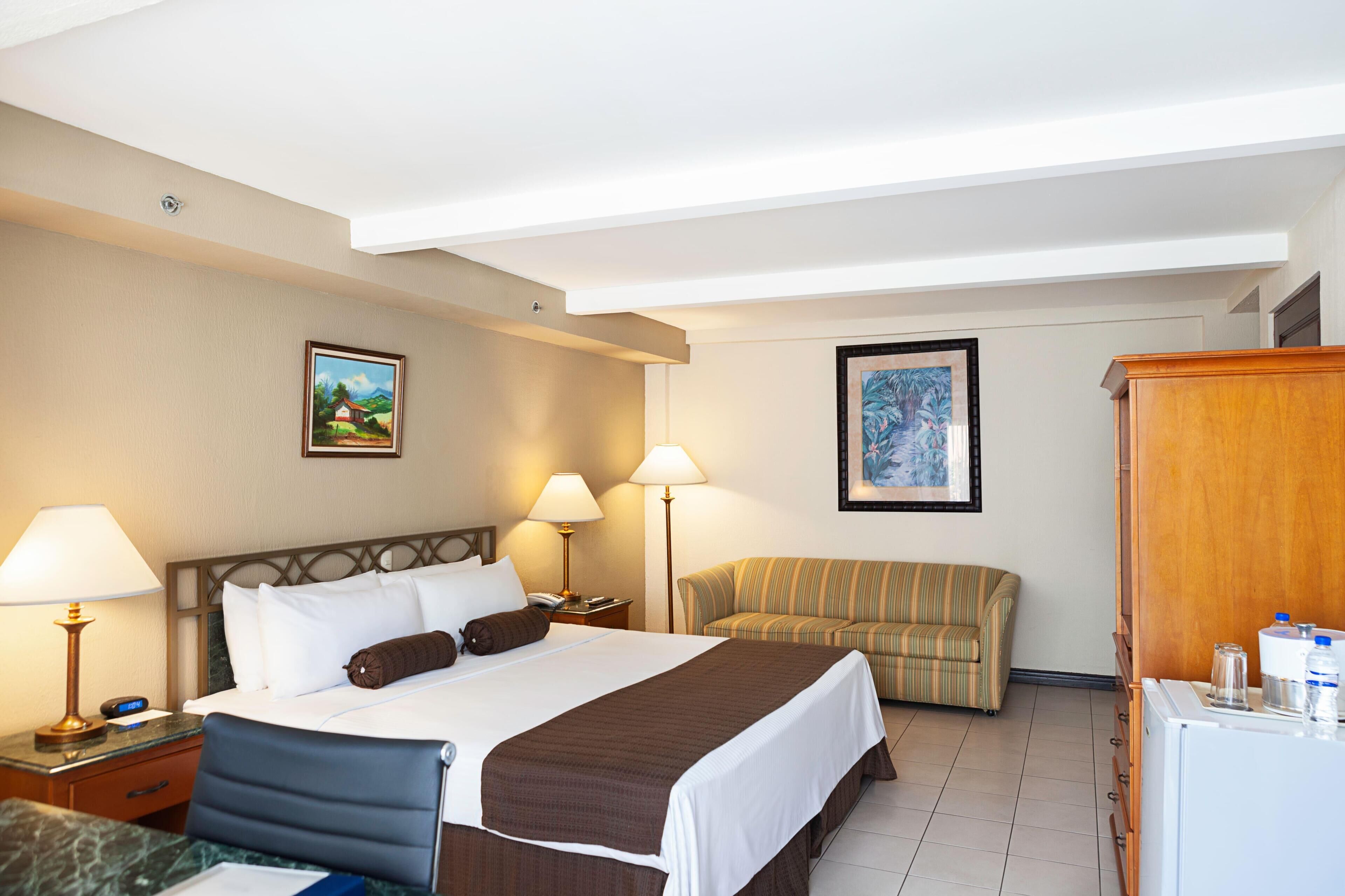 A hotel room with a bed, two bedside lamps, a sofa, a desk with a lamp, and a refrigerator.