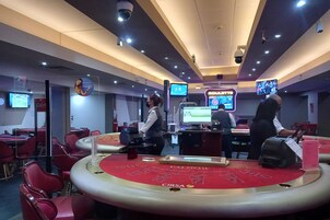 Un salón de casino con mesas de blackjack, cámaras de seguridad y pantallas digitales mostrando juegos.