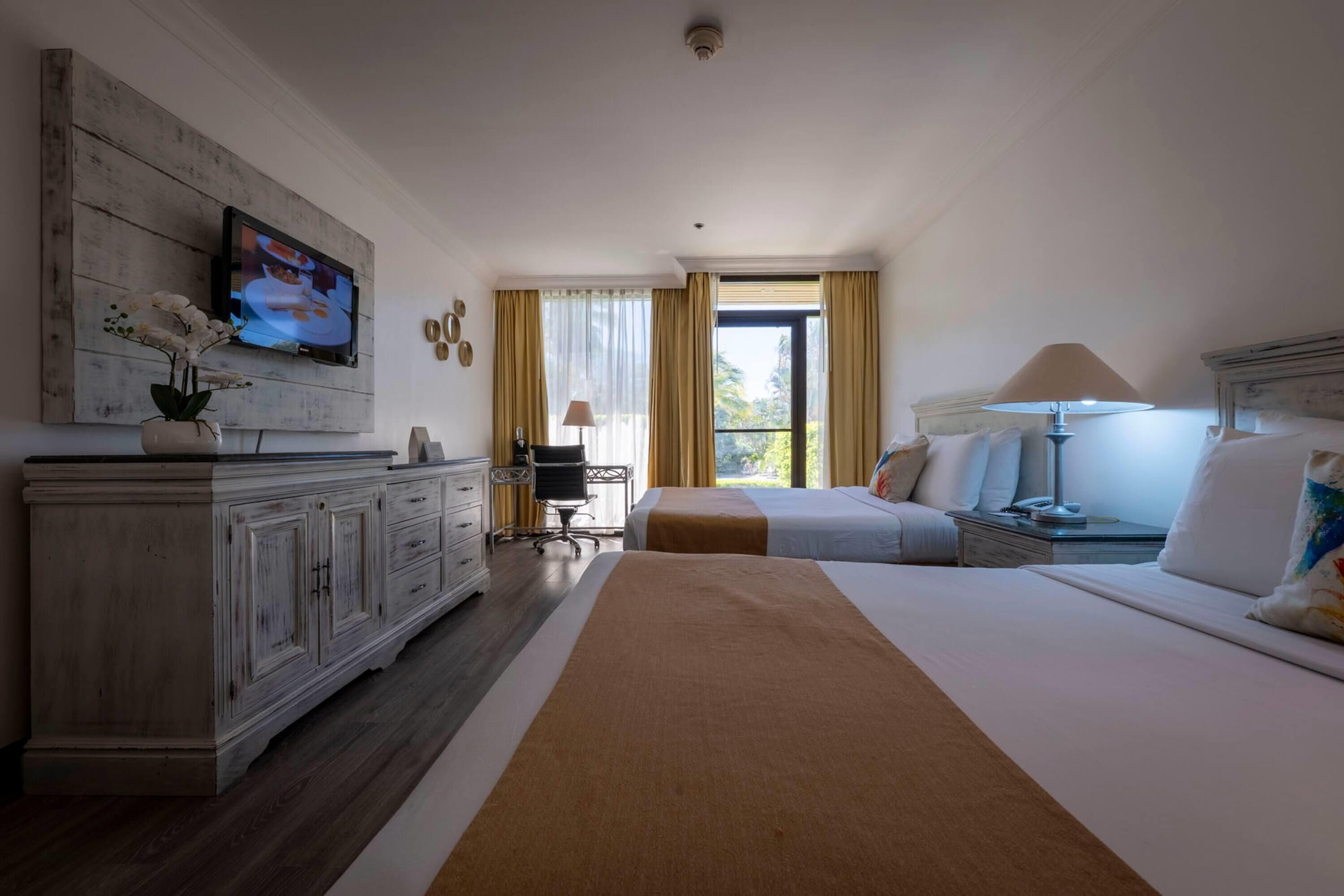 A hotel room with a large bed, a desk, a television mounted on a wooden panel, and a window with curtains.