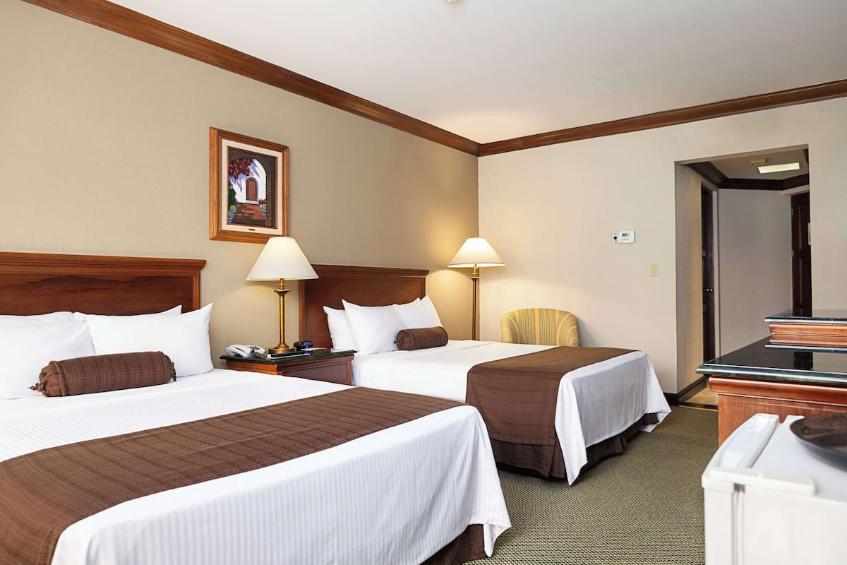A hotel room with two beds, a wooden headboard, a painting on the wall, and a desk with a lamp.