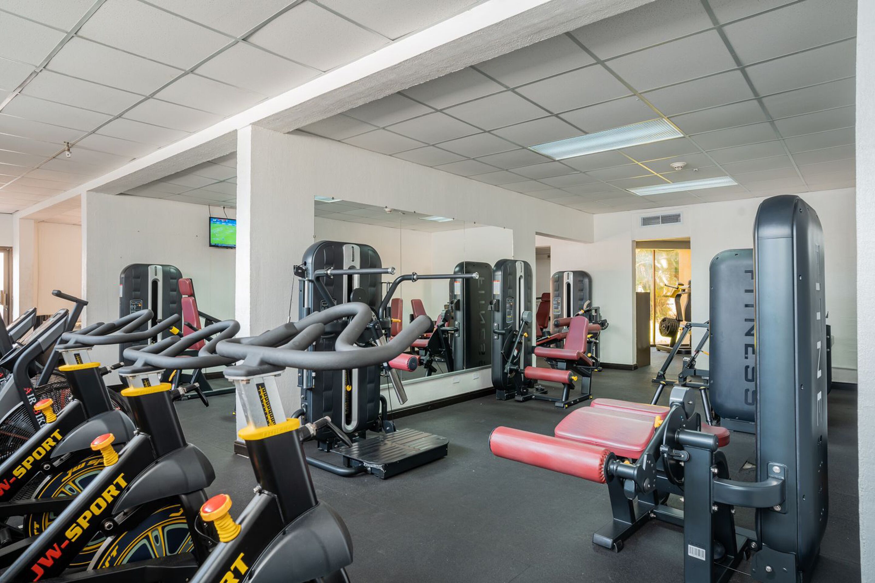 A gym with exercise equipment, mirrors, and a television on the wall.