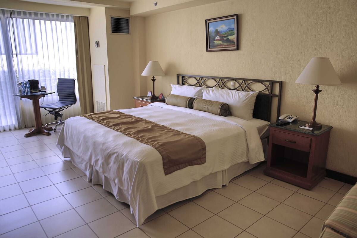 A hotel room with a large bed, two bedside tables with lamps, a chair, a desk, and a painting on the wall.