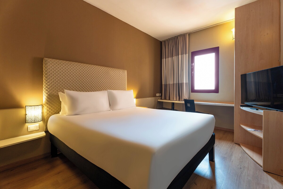 Une chambre d’hôtel avec un grand lit, une télévision, un bureau et une fenêtre avec des rideaux.