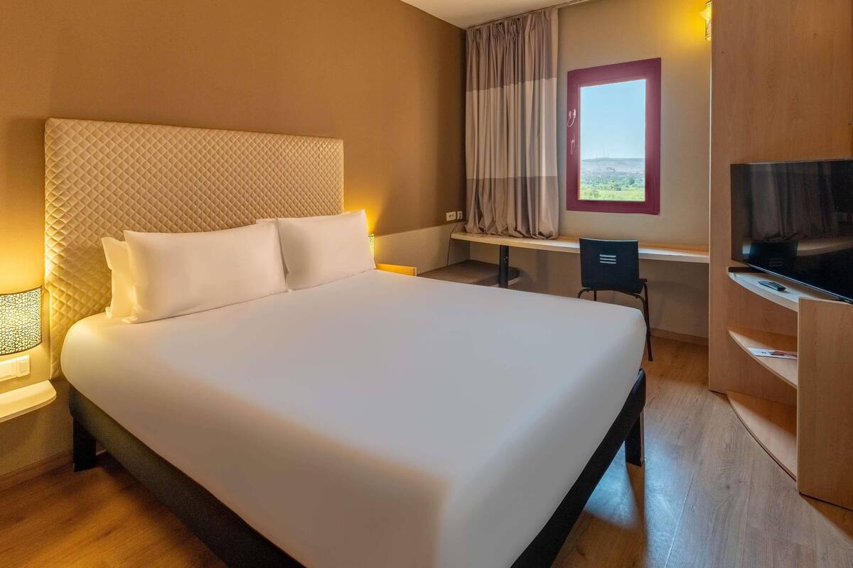 Une chambre d’hôtel avec un grand lit, un bureau, une chaise, une télévision et une fenêtre avec des rideaux.