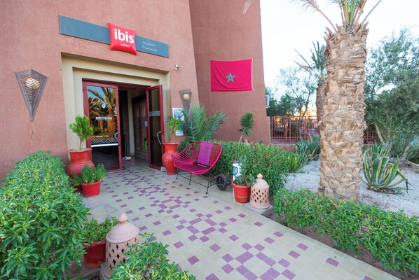 Exterior de un hotel ibis con suelo de baldosas a cuadros, plantas en macetas, una palmera y una bandera marroquí.