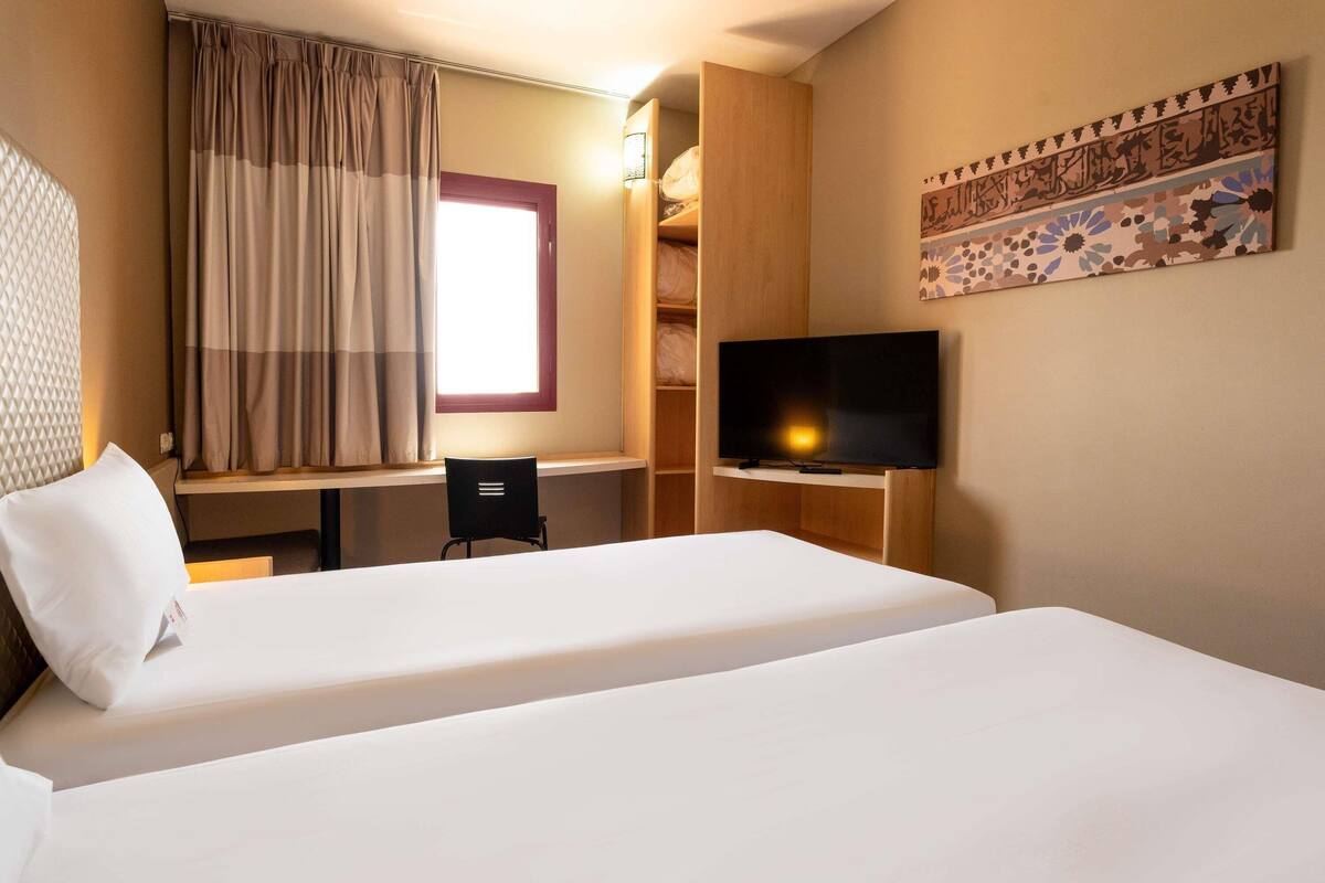 Une chambre d’hôtel avec deux lits, un bureau, une chaise, une télévision et une fenêtre avec des rideaux.