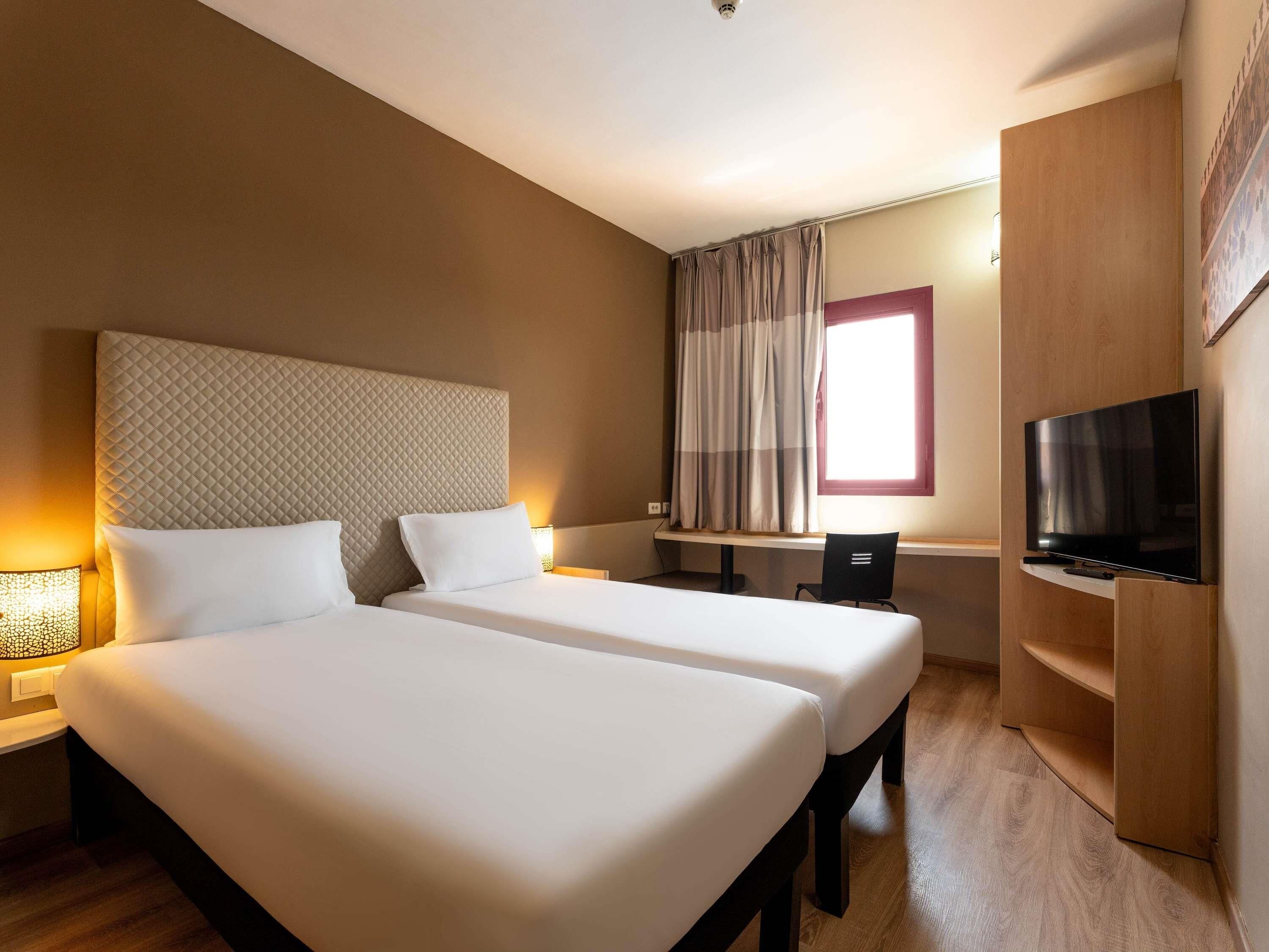 A hotel room with two beds, a desk, a TV, and a window with curtains.