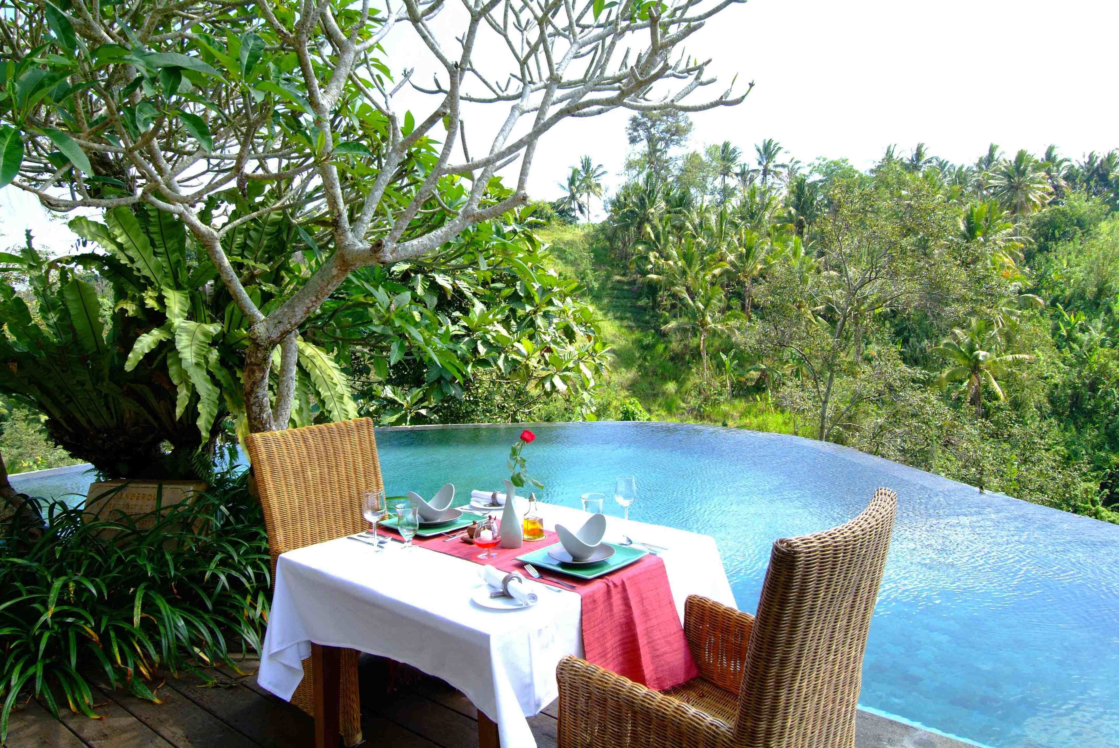 A dining table with a white tablecloth and red napkins, set against a lush backdrop of greenery and a clear blue pool.