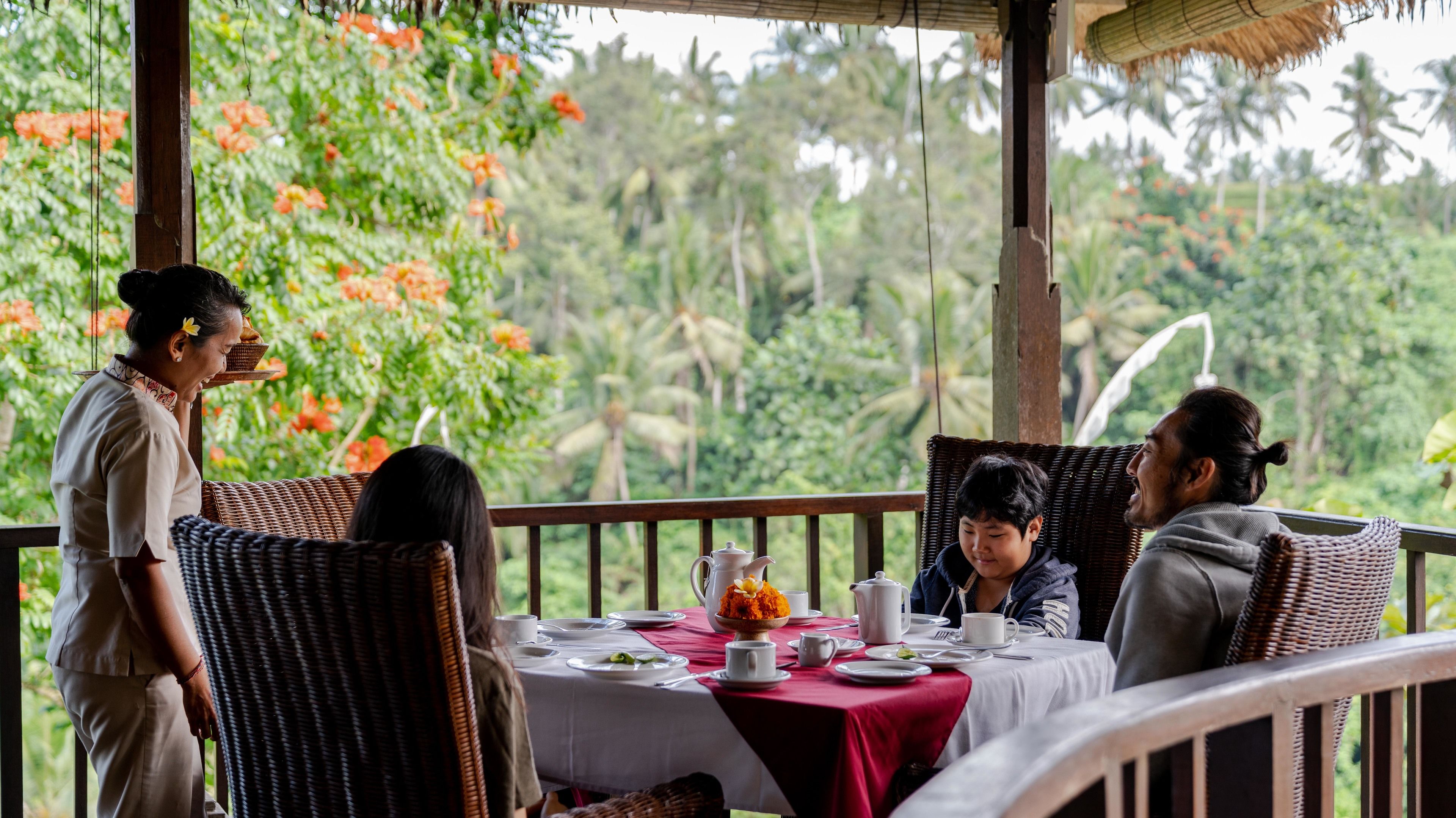 Un coin repas avec une table dressée pour quatre personnes, entourée de chaises en osier, offrant une vue sur une végétation luxuriante et des palmiers.