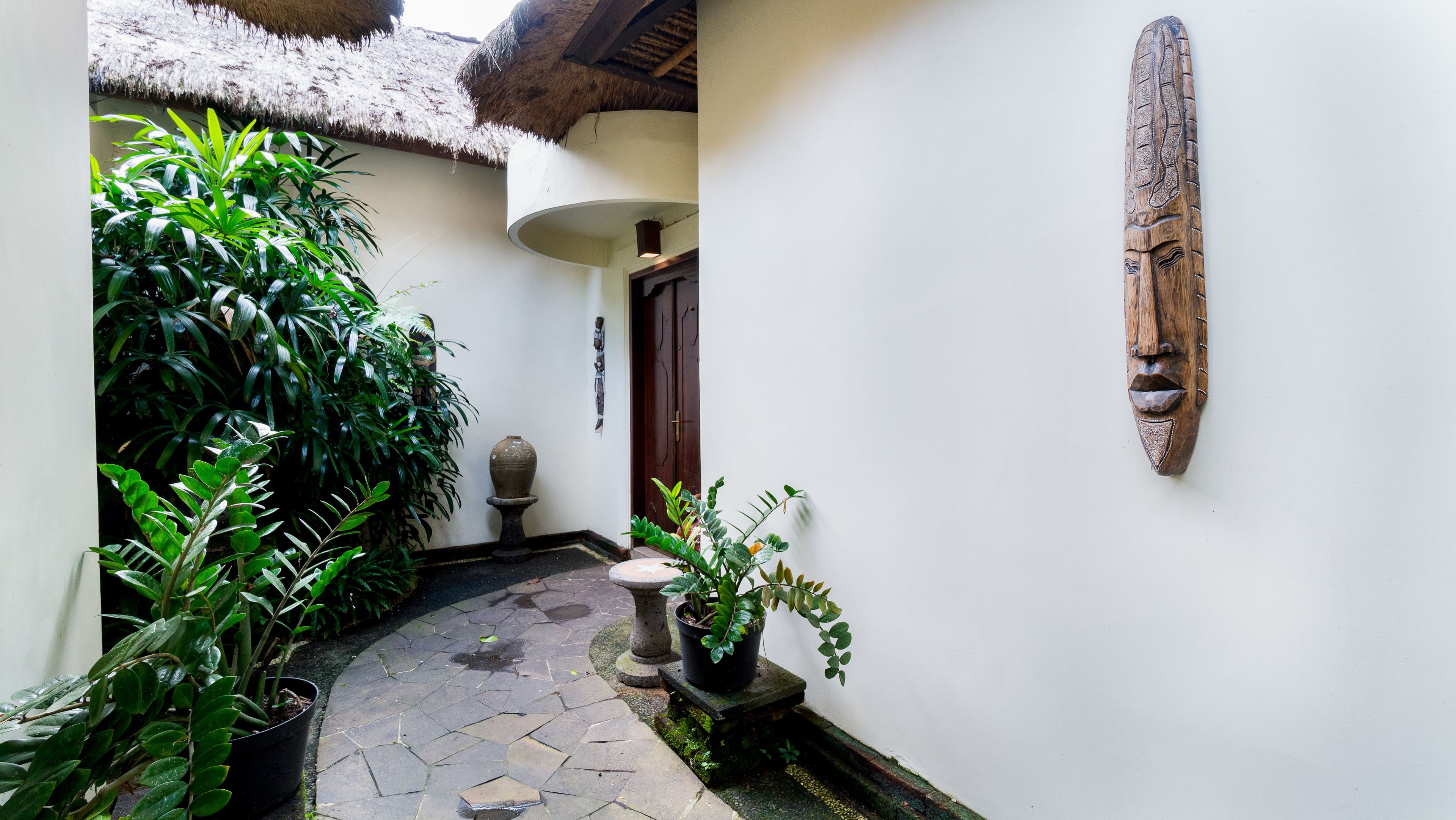 A narrow courtyard with a thatched roof, potted plants, a stone pathway, and a wooden door.