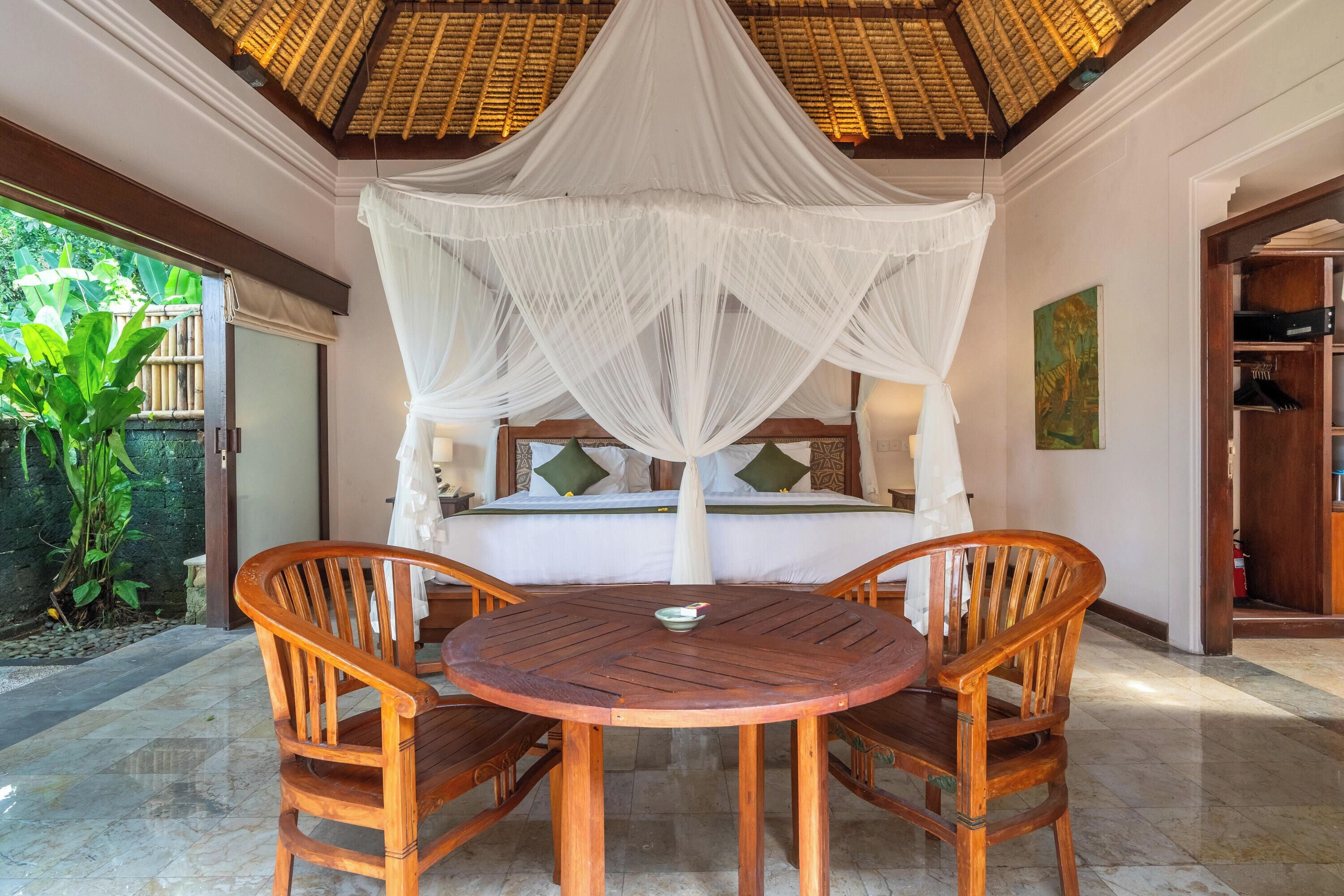 A room with a bed under a canopy, a round wooden table with chairs, and a view of greenery outside.