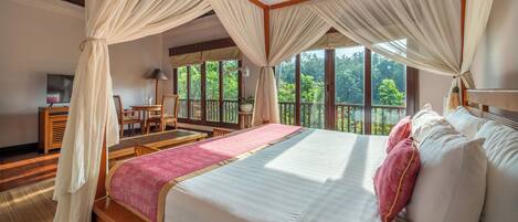 A spacious bedroom with a large bed, a wooden headboard, a small desk, and a chair. The room has large windows offering a view of greenery outside.