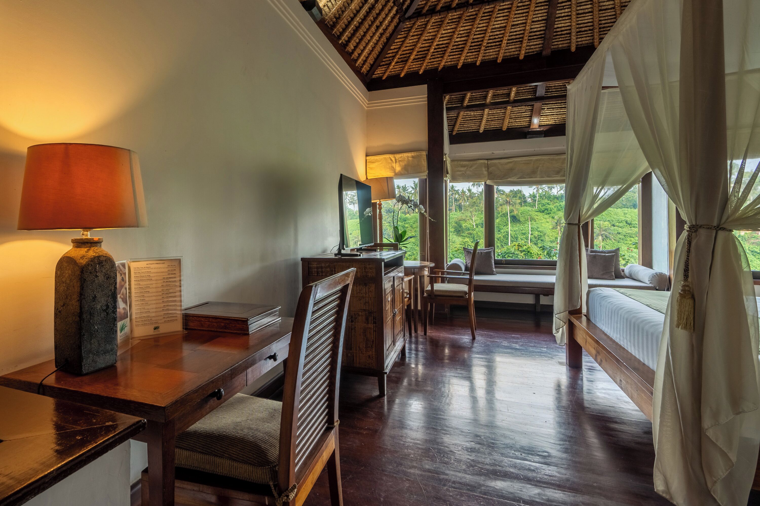 A room with a wooden desk, a lamp, a bed with a canopy, a window with a view of greenery, and a bench by the window.