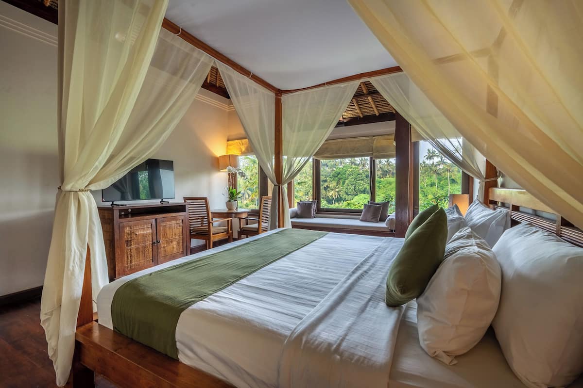Une chambre à coucher comprenant un lit, un bureau, une chaise et donnant sur un paysage verdoyant grâce à de grandes fenêtres.