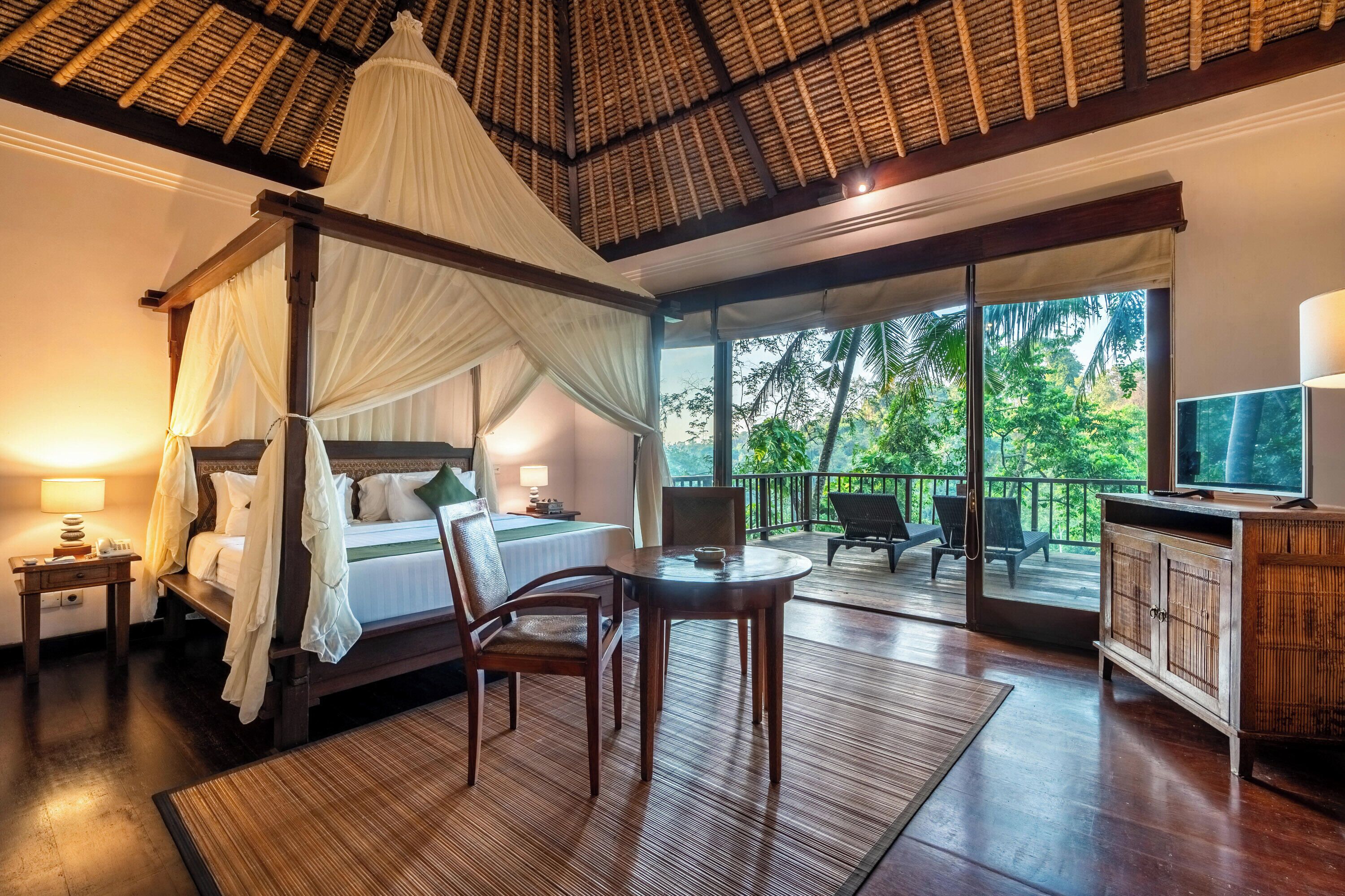 A four-poster bed with a canopy, a television, a dining table, and a balcony with a view of greenery.