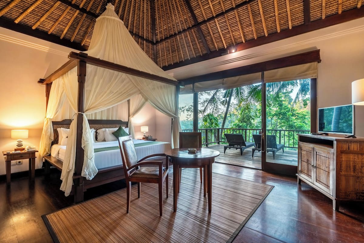 A four-poster bed with a canopy, a television, a dining table, and a balcony with a view of greenery.
