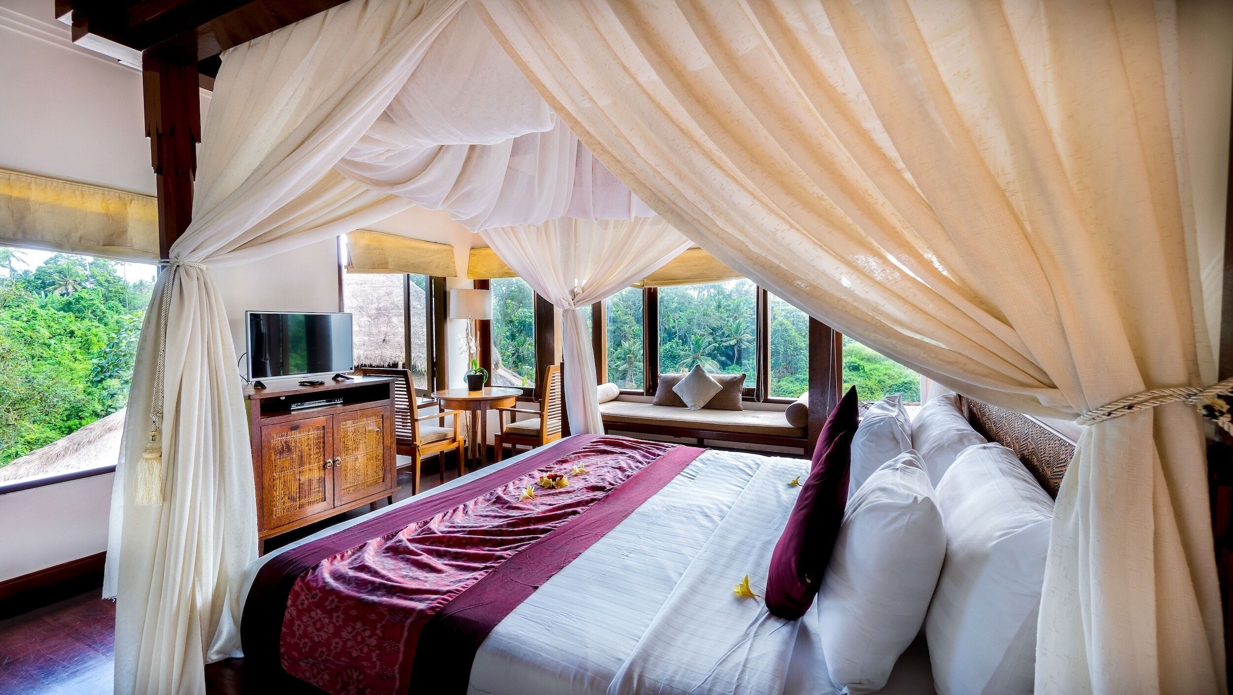 A bedroom with a canopy bed, a television, a desk, and a view of greenery.