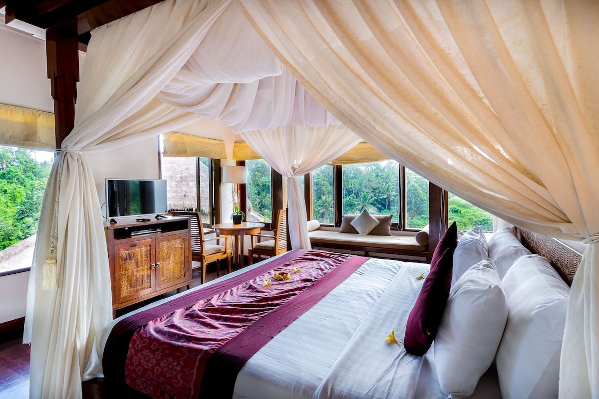 A bedroom with a canopy bed, a television, a desk, and a view of greenery.
