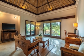 A spacious living room with a large TV, wooden furniture, a ceiling fan, and large windows offering a view of greenery.