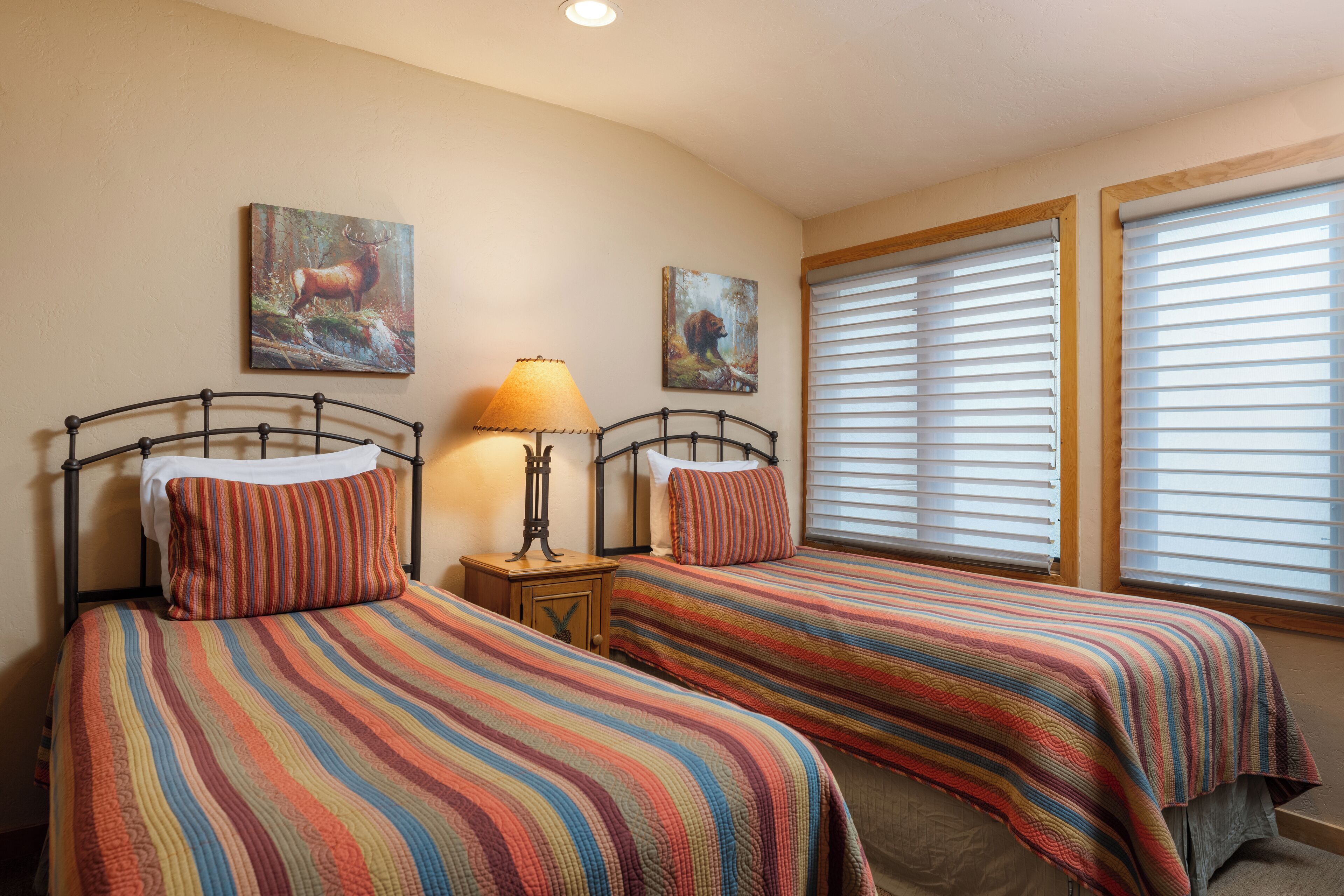 A bedroom with two beds, striped bedding, a nightstand with a lamp, and two wall-mounted paintings.
