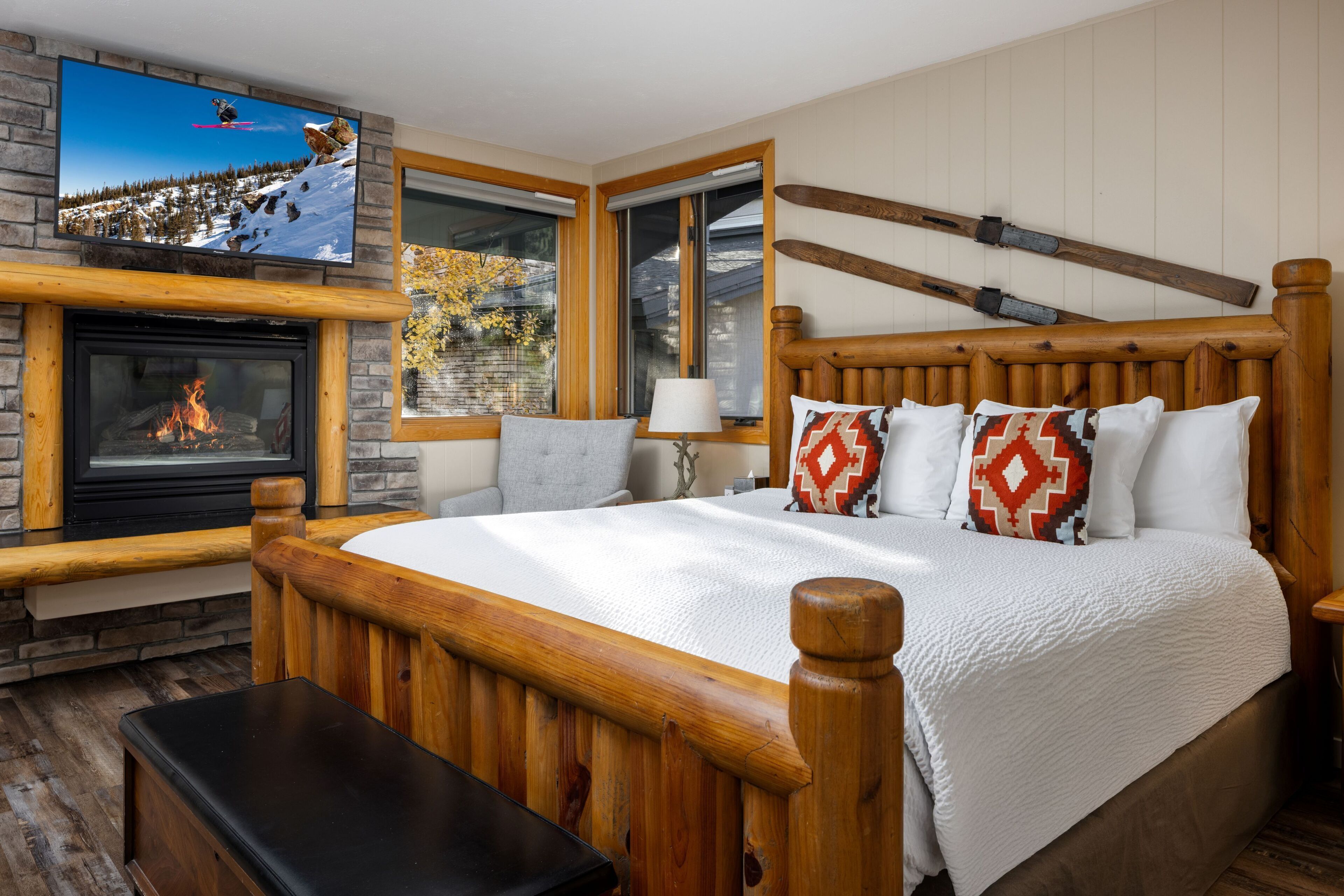 Une chambre dotée d’un grand lit, d’une tête de lit en bois, d’une cheminée, d’un téléviseur fixé au-dessus de la cheminée et d’une fenêtre donnant sur la forêt.