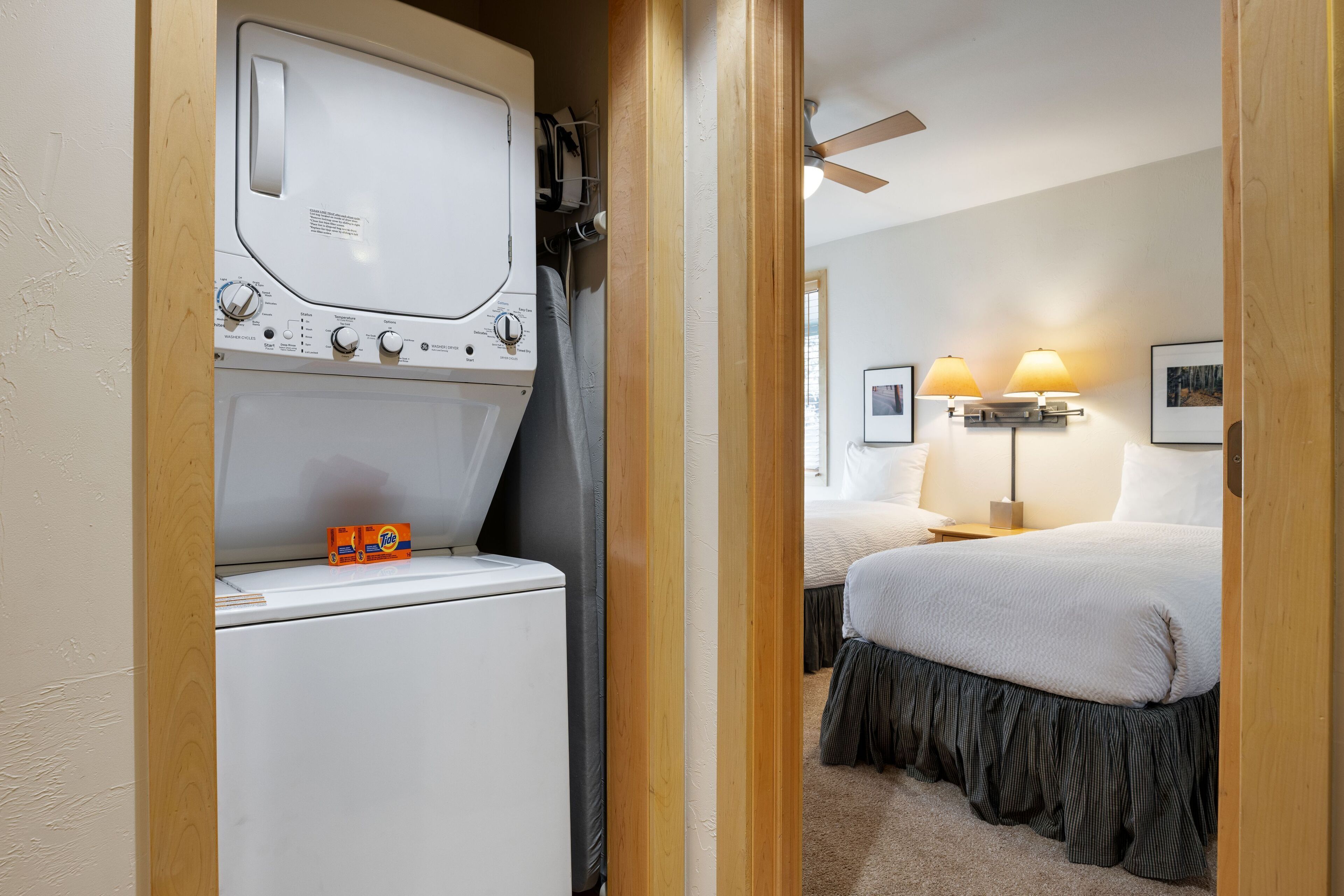 Ein Hotelzimmer mit Waschmaschine, einem Bett mit dunklem Bettdeckenrock, zwei Nachttischlampen und einem Deckenventilator.