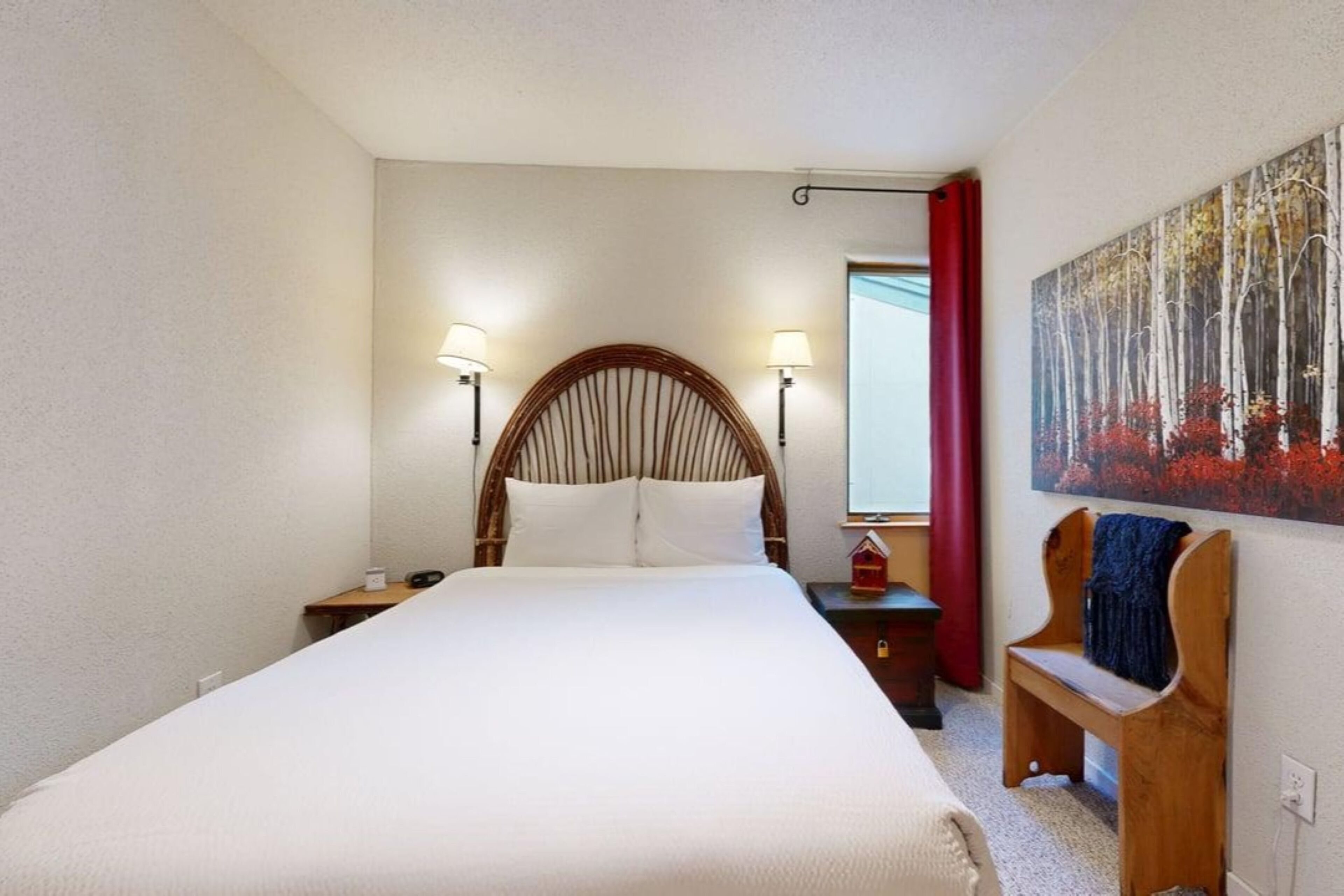 Ein Schlafzimmer mit einem großen Bett, zwei Nachttischlampen, einer Holzbänkl, einem Gemälde und einem Fenster mit roten Vorhängen.
