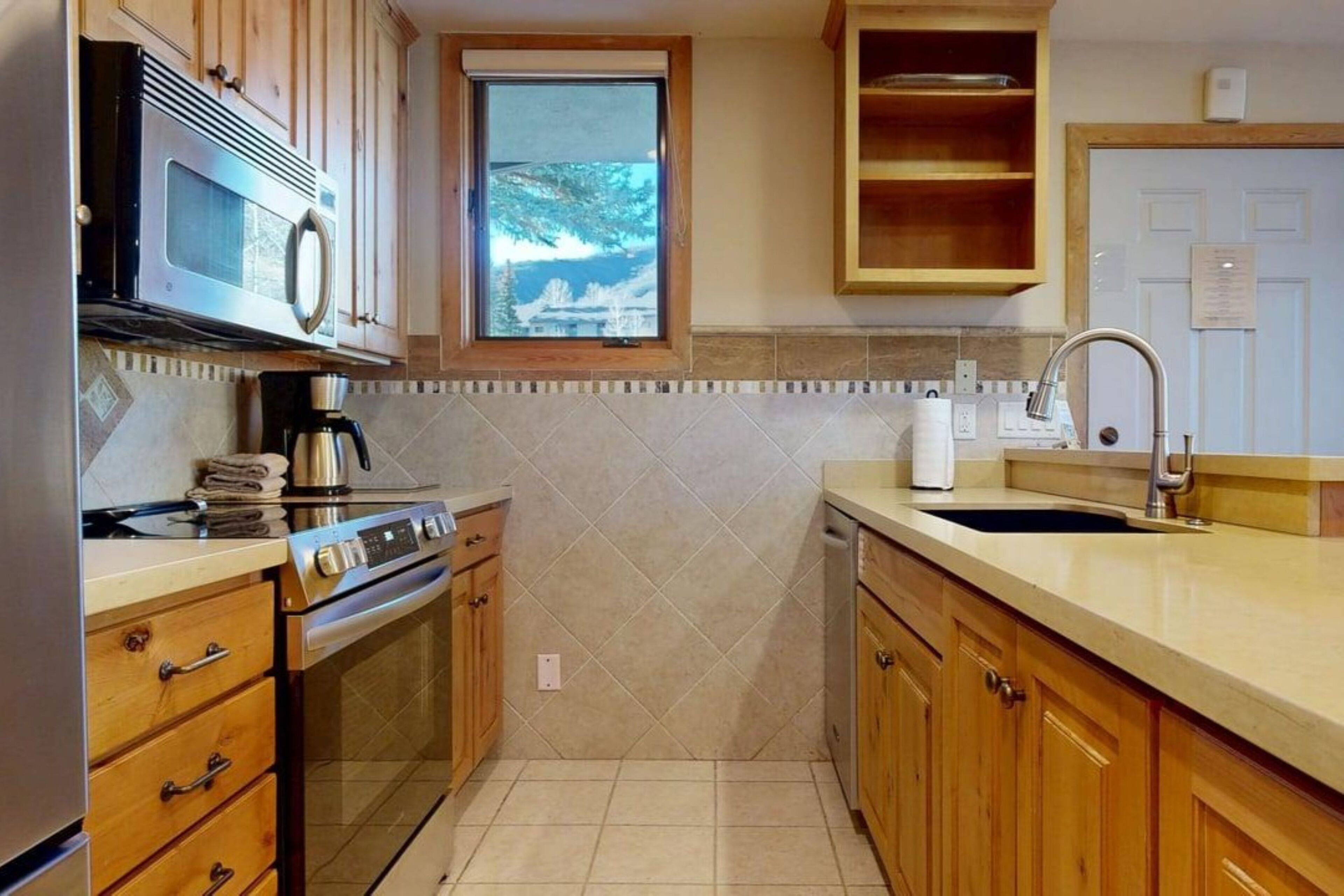 Una cocina con armarios de madera, microondas, estufa, fregadero y ventana con vistas a árboles.
