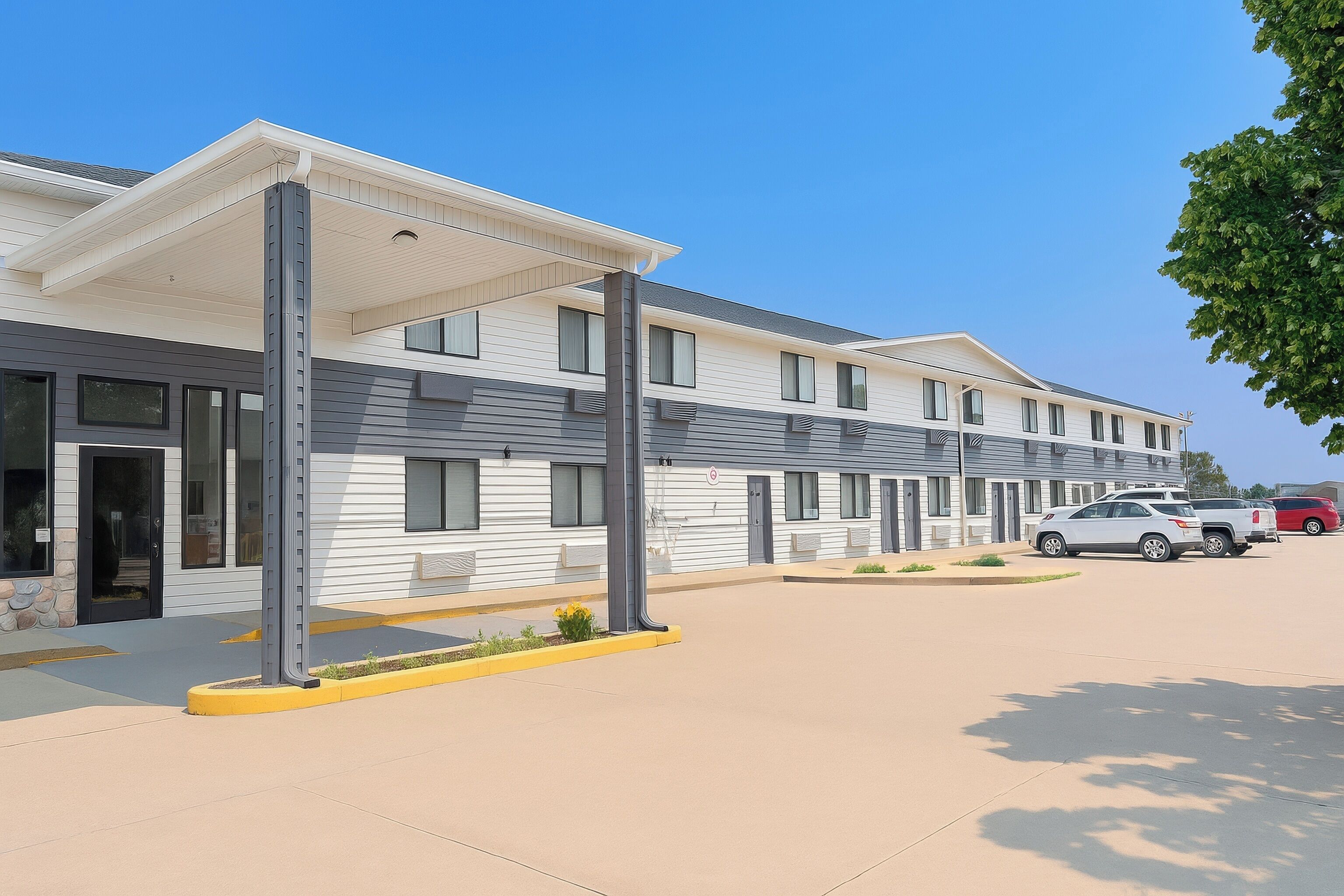 A modern hotel with a covered entrance, multiple windows, and a parking area with cars.